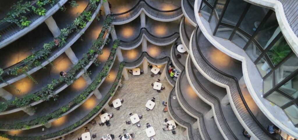 Aerial view of a modern multi-level atrium with green plants, round terraces, and small tables, creating a vibrant social space.